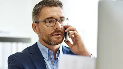 A businessman in a suit and glasses talks on his phone while reviewing a document at his desk.