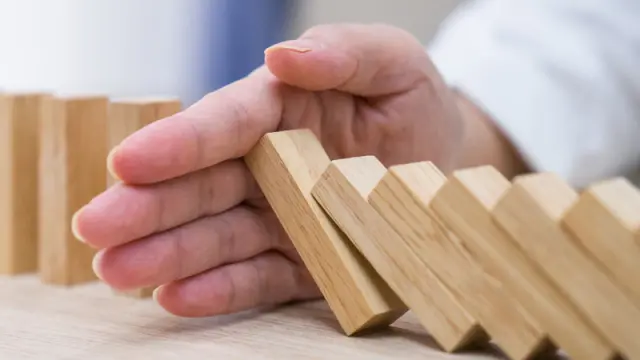 A hand intervenes to stop a chain reaction of falling wooden dominoes on a table.