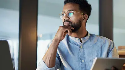 A thoughtful man with glasses and a beard holds a tablet and rests his chin on his hand.