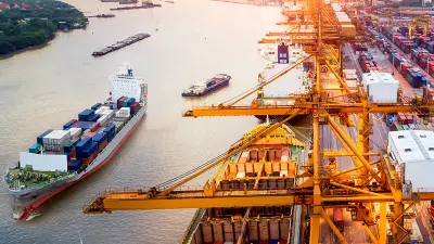Aerial view of a busy shipping port at sunset, with cranes loading cargo ships and a city skyline in the background.