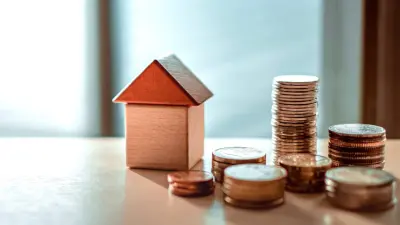 A small wooden block house sits on a table next to several stacks of coins of varying heights.