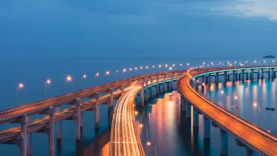 A long exposure shot of a brightly lit, curving highway bridge over the ocean at dusk, showing light trails from traffic.