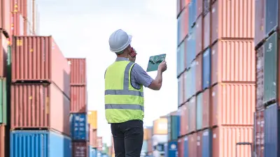 A worker in a hard hat and safety vest stands between stacks of shipping containers, using a walkie-talkie and a tablet.