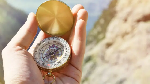 A person's hand holds a brass compass, with a blurry, sunny mountain landscape in the background.