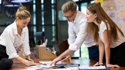 Three colleagues collaborate in a modern office, gathered around a wooden table reviewing charts and documents.