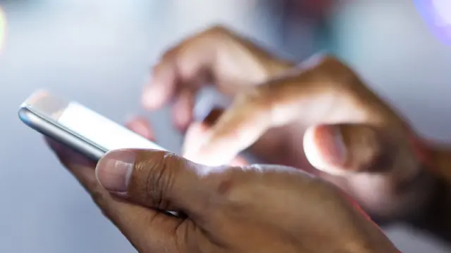 Close-up of hands holding and using a smartphone with a brightly lit screen at night, with city lights blurred in the background.