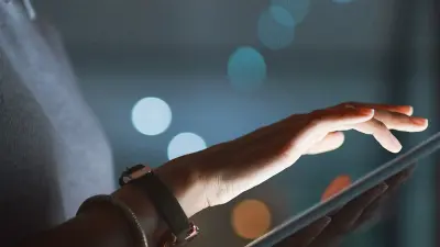 A woman's hand scrolls on a glowing tablet screen at night, with out-of-focus lights in the background.