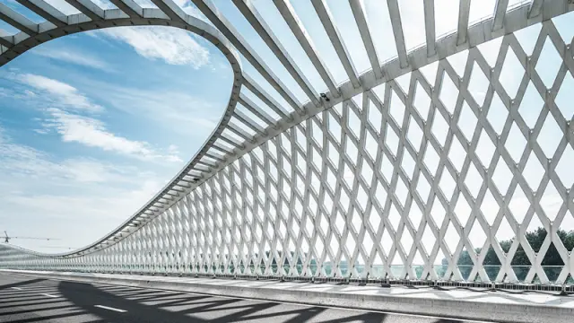 An empty road on a modern bridge with a white, geometric, wave-like canopy casting shadows on the asphalt.