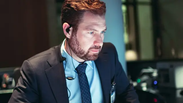 A man in a suit with an earpiece looks intently at a computer screen in a dimly lit control room.