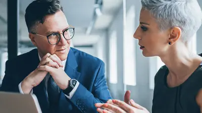 A man in a suit and glasses listens intently as a woman with short gray hair speaks to him across a table in an office.