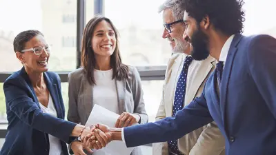 A diverse group of four smiling business professionals shake hands in front of a large window.