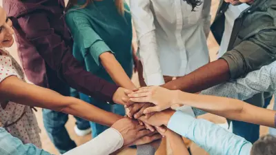 A diverse group of smiling people stand in a circle and stack their hands together in the center.